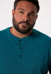 Tyrkysová bavlněná košile s kulatým výstřihem, tříknoflíkovou légou a embosovaným logem na levé části hrudi. Měkká textura.