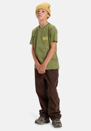 Green cotton t-shirt with short sleeves and a logo print, paired with brown corduroy pants and a yellow knit beanie. Footwear features grey accents.