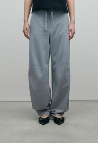 Pantalons larges gris en tissu texturé, avec une taille à cordon de serrage et des détails de couture subtils, assortis à des chaussures noires à bouts pointus.