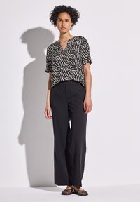 Schwarz und braun gemustertes, kurzärmliges Oberteil, lockere Passform, kombiniert mit weiten schwarzen Hosen und flachen schwarzen Netzschuhen. Minimalistisches Design, lässiger Stil.