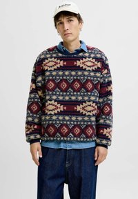 Fleece trui met textuur in rode, marineblauwe en crèmewitte geometrische patronen, met een ronde hals en een oversized pasvorm, over een denim shirt gedragen.