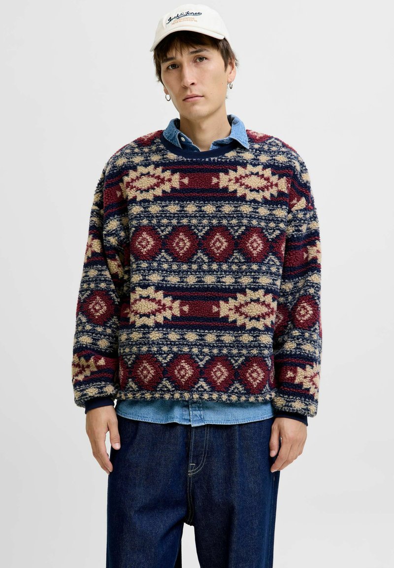 Fleece trui met textuur in rode, marineblauwe en crèmewitte geometrische patronen, met een ronde hals en een oversized pasvorm, over een denim shirt gedragen.