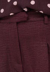 Gros plan sur un pantalon en tissu bordeaux avec une ceinture et des passants, associé à un chemisier à pois bordeaux rentré.