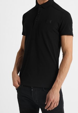 Mann trägt eng anliegendes schwarzes kurzärmeliges Poloshirt mit Knöpfen und schwarze Jeans, steht mit einer Hand in der Hosentasche vor weißem Hintergrund.