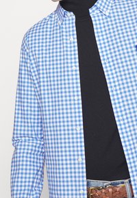 Blå och vit rutig skjorta med nedknäppt krage, långa ärmar och en svart polo tröja under, kombinerad med ett vävt brunt bälte.