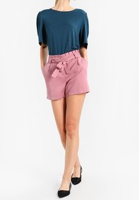 Top corto a maniche corte color teal con spalle arricciate, abbinato a pantaloni corti rosa a vita alta con cintura annodata e tasche laterali. Scarpe nere.