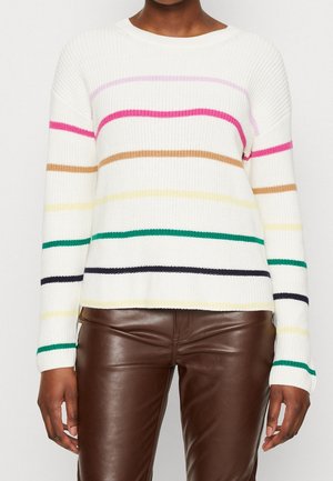 Personne portant un pull en tricot blanc avec des rayures horizontales multicolores et un pantalon en cuir marron, se tenant devant un fond uni.