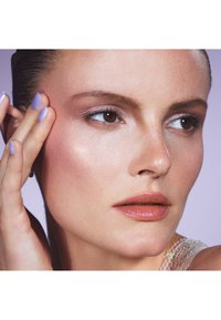 Primo piano di un volto con pelle liscia, occhi nocciola chiari e rossetto nude. Unghie viola accentuano l'aspetto naturale. L'illuminazione soffusa mette in risalto i dettagli.
