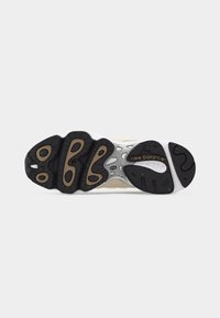 Semelle en caoutchouc noire et marron d'une chaussure New Balance avec un motif de bande de roulement et le logo "new balance" sur une base grise et blanche.