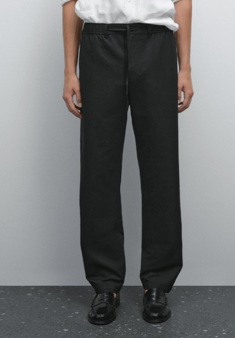 Person wearing black straight-leg trousers, black slip-on shoes, and a white tucked-in shirt standing against a plain background.