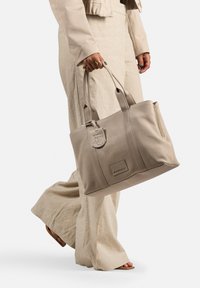 Beige leren schoudertas met dubbele handvatten, een ritszak, reliëf logo-details en een gestructureerde vorm. Gecombineerd met lichtlinnen broeken.