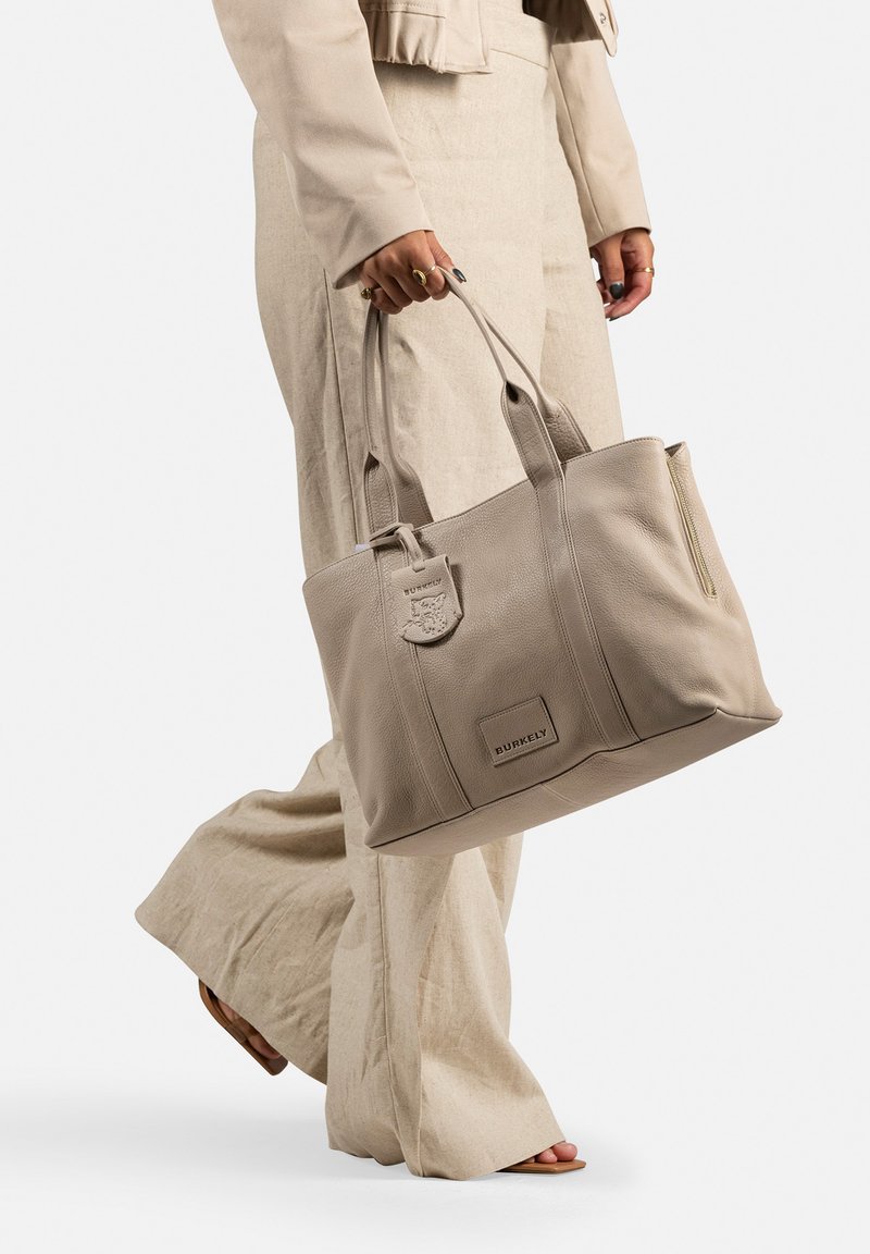 Beige leren schoudertas met dubbele handvatten, een ritszak, reliëf logo-details en een gestructureerde vorm. Gecombineerd met lichtlinnen broeken.