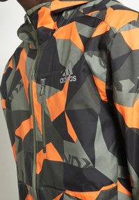 Giacca camouflage in arancione, nero e verde con motivi geometrici. Dotata di cappuccio, zip e logo Adidas sul petto.