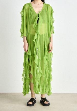 Mujer con un vestido largo, transparente y verde lima con volantes y lazo delantero, y sandalias negras, de pie frente a un fondo claro y sencillo.