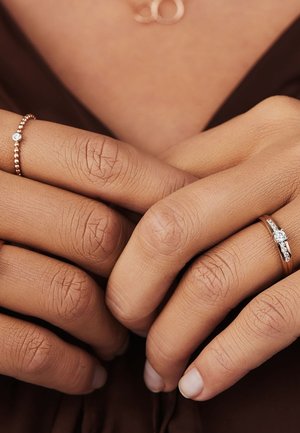 Zwei Ringe an den Händen: Der eine ist ein dünner Roségoldring mit kleinen Perlen, der andere hat einen zentralen Stein und kleinere Akzente aus Roségold.
