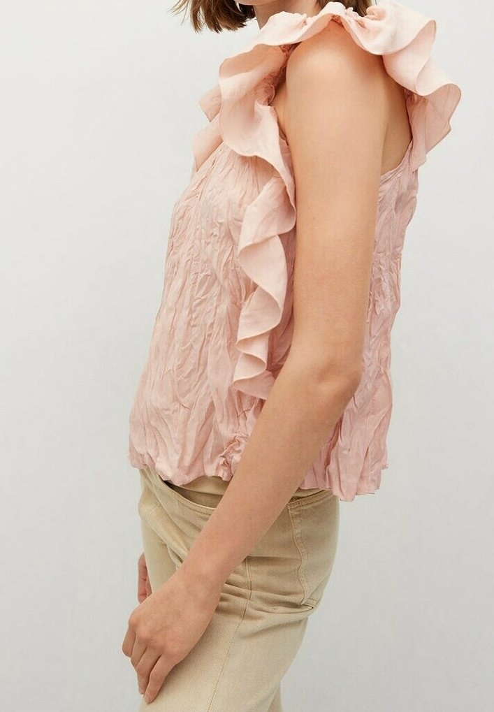 Vaaleanpunainen hihaton paita, jossa on röyhelökaulus, ryppyinen rakenne ja rypistetty helma. Yhdistetty vaaleisiin housuihin, nähty sivulta.