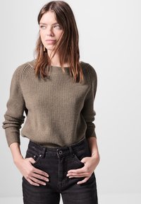 Maglione a coste verde oliva con scollo a barca, abbinato a jeans neri a vita alta. Mani appoggiate sui fianchi, posizione rilassata.