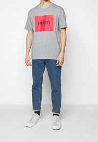 Camiseta de algodón gris con un gran cuadrado rojo que lleva la palabra "HUGO" en texto negro. Acompañada de jeans de mezclilla azul y zapatillas blancas.