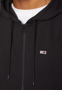 Svart zip-up hoodie i mjukt material, med ett vitt och färgglatt logomärke på bröstet och dragsnören med profilerade spetsar.