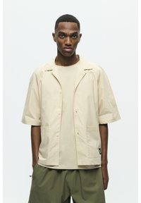 Beige Jacke mit kurzen Ärmeln, mit Reverskragen, vorn mit Knöpfen und zwei Taschen. Getragen über einem passenden beigen Hemd. Olivgrüne Hose.