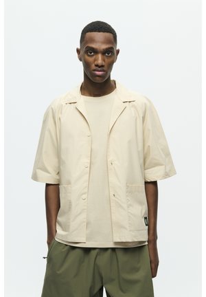 Short-sleeve beige jacket with a notched collar, front buttons, and two pockets. Worn over a matching beige shirt. Olive green pants.