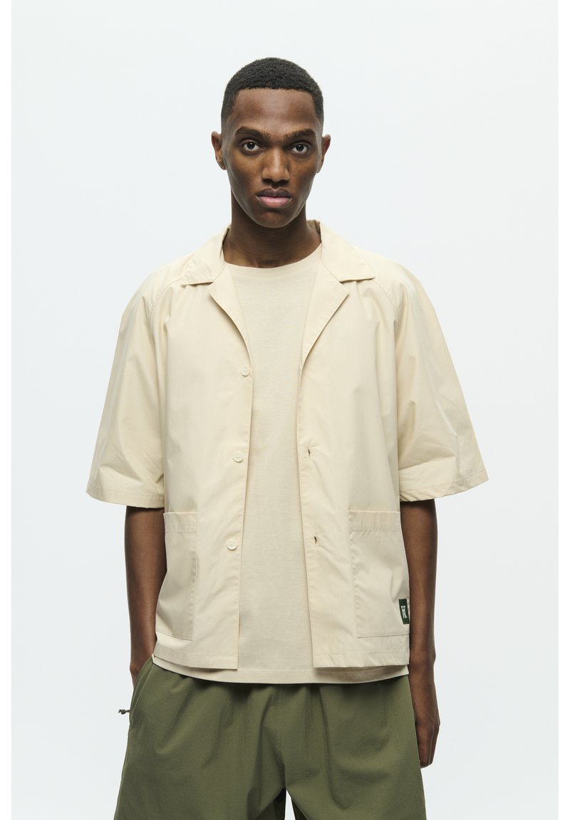 Beige Jacke mit kurzen Ärmeln, mit Reverskragen, vorn mit Knöpfen und zwei Taschen. Getragen über einem passenden beigen Hemd. Olivgrüne Hose.