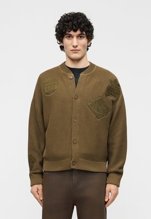 Fiatal férfi, barna texturált gombos kardigánban, hímzett foltokkal, sötétbarna nadrággal, egyszínű háttér előtt állva.