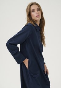 Rochie button-up bleumarin din țesătură moale, cu mâneci lungi, guler și buzunare laterale. Textură netedă, cu o croială lejeră.