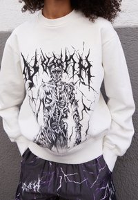 Vitt sweatshirt med en svart grafisk tryck av en skelettfigur med intrikata detaljer. Återfinns med mönstrade svarta och lila byxor.