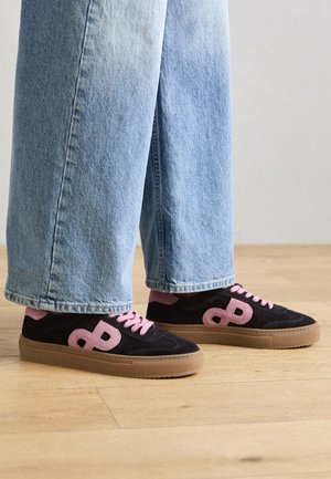 Osoba nosząca jasnoniebieskie szerokie jeansy i czarne trampki z różowymi sznurowadłami, logo i beżowymi podeszwami, stojąca na drewnianej podłodze.