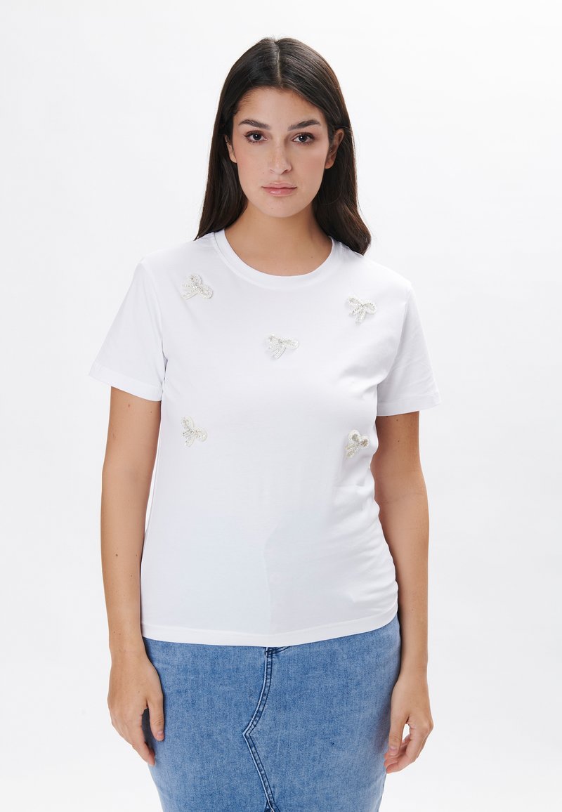 Camiseta blanca de manga corta hecha de algodón suave, con detalles de lazo plateado bordados en el frente. Combinada con una falda de mezclilla.