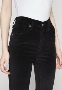 Svarta kordbyxor med en figursydd design, hög midja, framfickor och detaljer i metall. Slät textur med subtila ribbor.