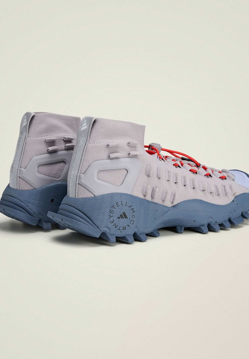 Scarpe sportive grigio chiaro con una tomaia testurizzata, lacci rossi e una suola in gomma blu con audaci modelli di trazione.