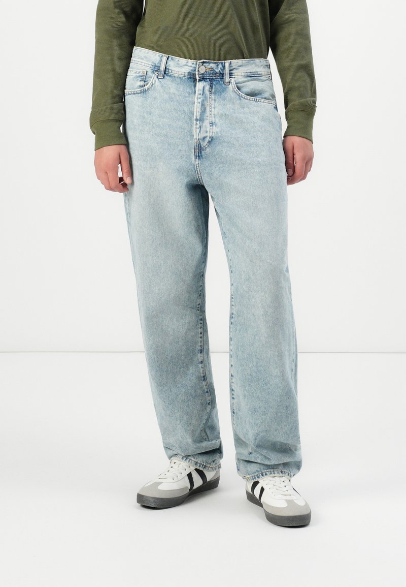Person som har på seg lyseblå, løse jeans, hvite og grå sneakers med svarte striper, og en olivengrønn langermet topp, stående på et hvitt gulv.