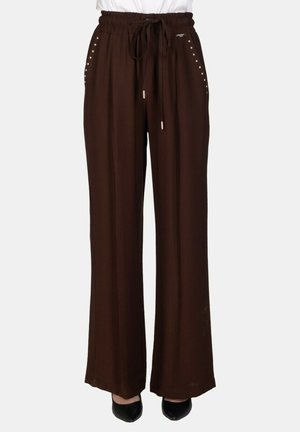 Pantaloni marroni a gamba larga con elastico in vita, coulisse, borchie argentate sulle tasche, indossati con scarpe nere appuntite e top bianco.