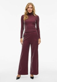 Ensemble bourgogne en deux pièces comprenant un turtleneck ajusté et un pantalon à jambes larges. Le tissu a un léger éclat. Chaussures à talons hauts noires.