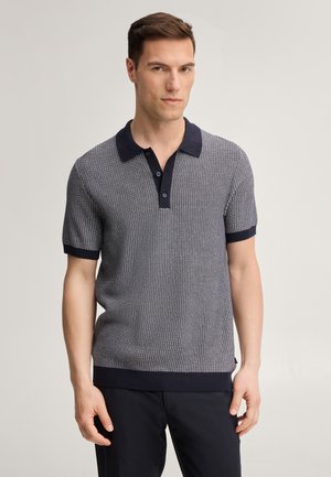 Uomo in piedi contro uno sfondo semplice, indossa una polo a maniche corte in maglia testurizzata con colletto e polsini scuri, e pantaloni scuri.