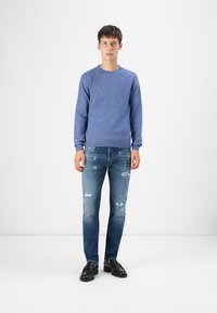 Blauwe gebreide trui met lange mouwen, geribde manchetten, gecombineerd met versleten blauwe jeans en zwarte leren schoenen. Effen achtergrond.