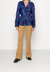 Osoba ubrana w wzorzystą fioletową bluzkę z bufkami i beżowe spodnie w stylu dzwonów, stojąca na białym tle.