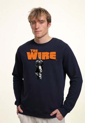 Granatowy sweatshirt z dużym pomarańczowym napisem "THE WIRE" oraz czarno-białą grafiką przedstawiającą osobę w płaszczu spacerującą. Miękka tkanina, luźny krój.