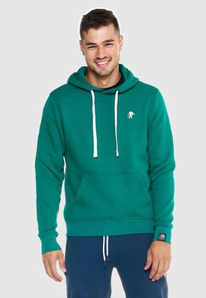 Lächelnder Mann mit grünem Hoodie mit weißen Kordeln und dunkelblauer Hose vor hellem, einfarbigem Hintergrund.