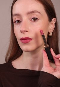 Rouge à lèvres rose mat dans un étui noir avec des accents dorés, tenu entre les doigts, sur un fond doux et avec un maquillage naturel sur les lèvres.