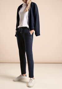 Marinblå bomberjacka med dragkedja, tillsammans med en vit topp med hög krage och matchande marinblå joggers. Vita sneakers fulländar looken.