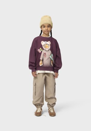 Kind met gevlochten haar, draagt een beige hoed, paarse trui met tijgerafbeelding, beige cargobroek en bruine sneakers, staand op een witte achtergrond.