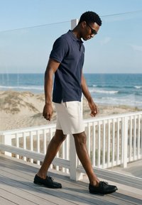 Homme portant des lunettes de soleil, un polo bleu marine, un short blanc et des chaussures noires, marchant sur une terrasse en bois près de la plage avec l'océan en arrière-plan.