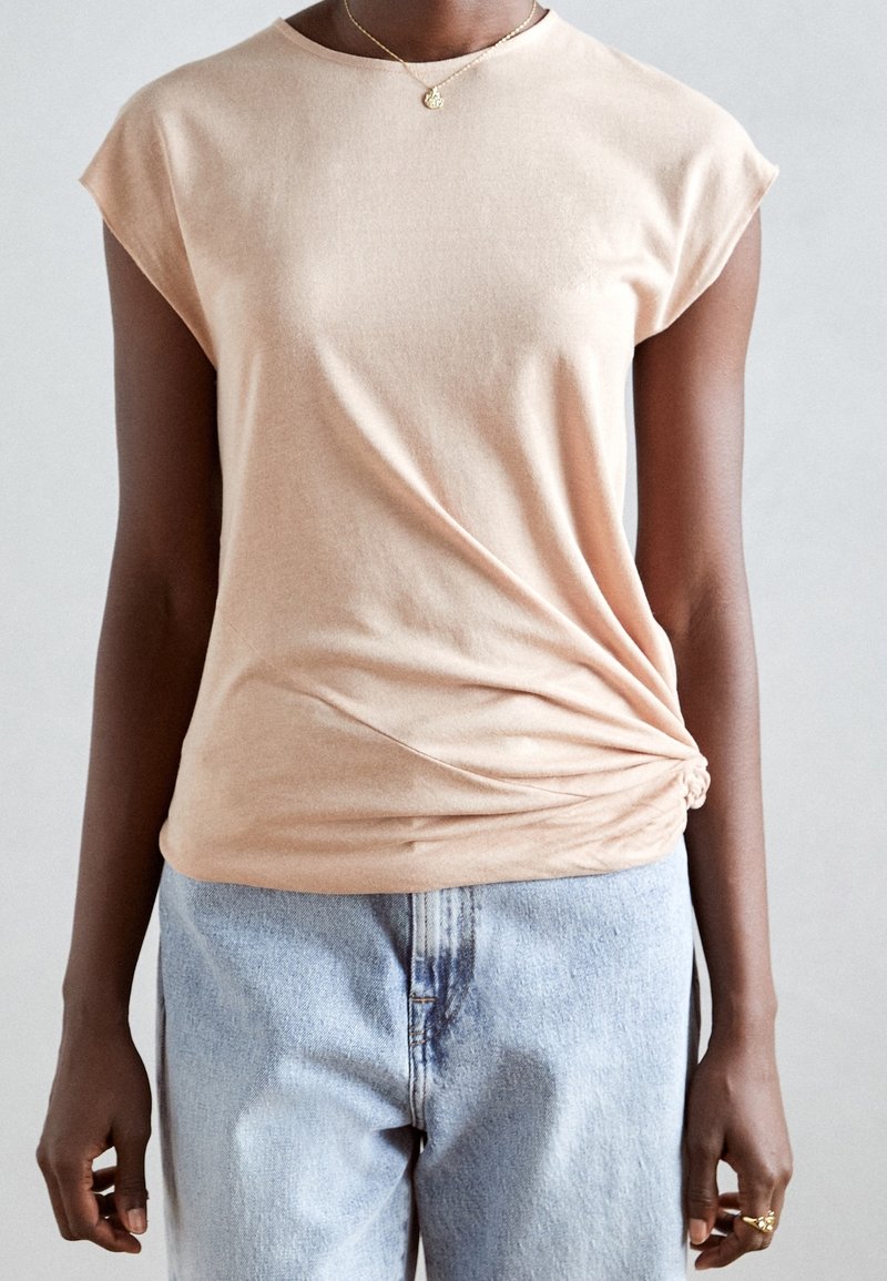 Camiseta de algodón suave de color durazno con mangas cortas, que presenta un detalle de nudo en el lado y un ajuste relajado, combinada con jeans azul claro.