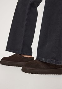 Zapatillas de ante marrón con un interior suave, que cuentan con una suela de goma oscura, gruesa y texturizada, y con el logo de la marca visible en el lateral.