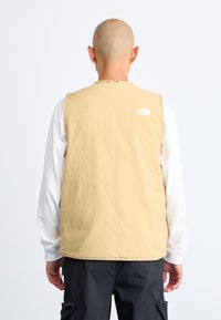 Beige vest made from lightweight fabric, featuring a round neck and a prominent white logo on the upper back. Worn over a long-sleeve white shirt.