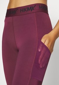 Bordowe legginsy z czarnym elastycznym paskiem w talii z logo "HKMX". Zawierają boczną kieszeń z przezroczystego materiału. Gładka tekstura.