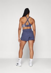 Sostén deportivo de color azul marino con diseño de espalda cruzada y tres franjas blancas; combinado con pantalones cortos de canalé azul marino. Modelo usa zapatillas y calcetas blancas.